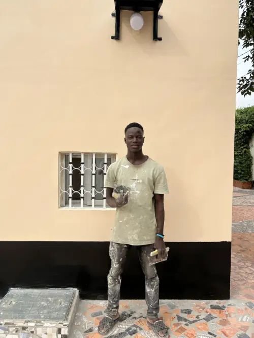 Young African painter and CEO of Pabi's Painting standing in front of a freshly painted house exterior in The Gambia, holding tools after a completed project.