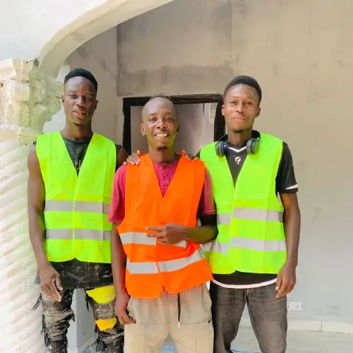 Three young African men from Pabi's Painting team in reflective vests, standing at a construction site, ready to begin a new painting project in The Gambia.